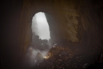 Son Doong Cave, Vietnam: View of Camp 1, our night camp. In the foreground a companion lights up towards a cloudy swath. In the background the light shines from the opening of a doline 300 meters deep to the ground. Dolines are natural shafts, which are caused by the collapse of the cave roof.