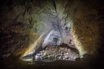 Blick durch den Mittelraum der Hang En Cave. Hier mischt sich das Licht unserer Höhlenlampen mit dem Tageslicht, welches durch den Ausgang der Höhle hereinströmt. Schon die Hang En ist riesig: Oben auf dem Schottenhaufen erkennt beim genauen Hinsehen das grüne Trikot eines Expeditionsmitglieds vor einem dunklen Quadrat im Hintergrund.

