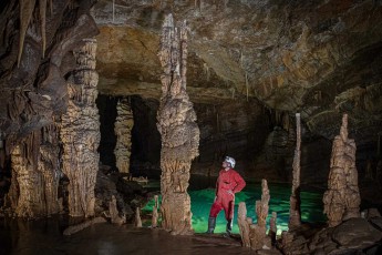 Gašper Modic stands between the monumental stalagmites of Križna Cave like a guardian of the underworld. The emerald green water at his feet reflects the ancient limestone formations, creating a scene of unreal beauty.