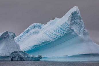 Gefrorene Wellen zeichnen sich auf der Flanke dieses Eisbergs ab – Spuren einstiger Schmelz- und Gefrierzyklen. Das tiefe Blau der Wasserlinie markiert den Bereich ständiger Erosion.