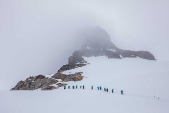 In Reih und Glied stapft eine Gruppe über das Schneefeld am Meusnier Point. Nebel verschluckt den Gipfel, nur dunkle Felsnasen durchbrechen das Weiß.