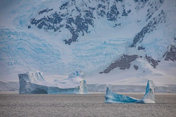 Zwei Eisberge in verschiedenen Verwitterungsstadien treiben vor der gewaltigen Gletscherlandschaft, jeder Eisberg erzählt seine eigene Geschichte aus Druck, Zeit und Zerfall. Beim rechten verrät das leuchtendes Blau einstmals verborgenes, uraltes Eis.